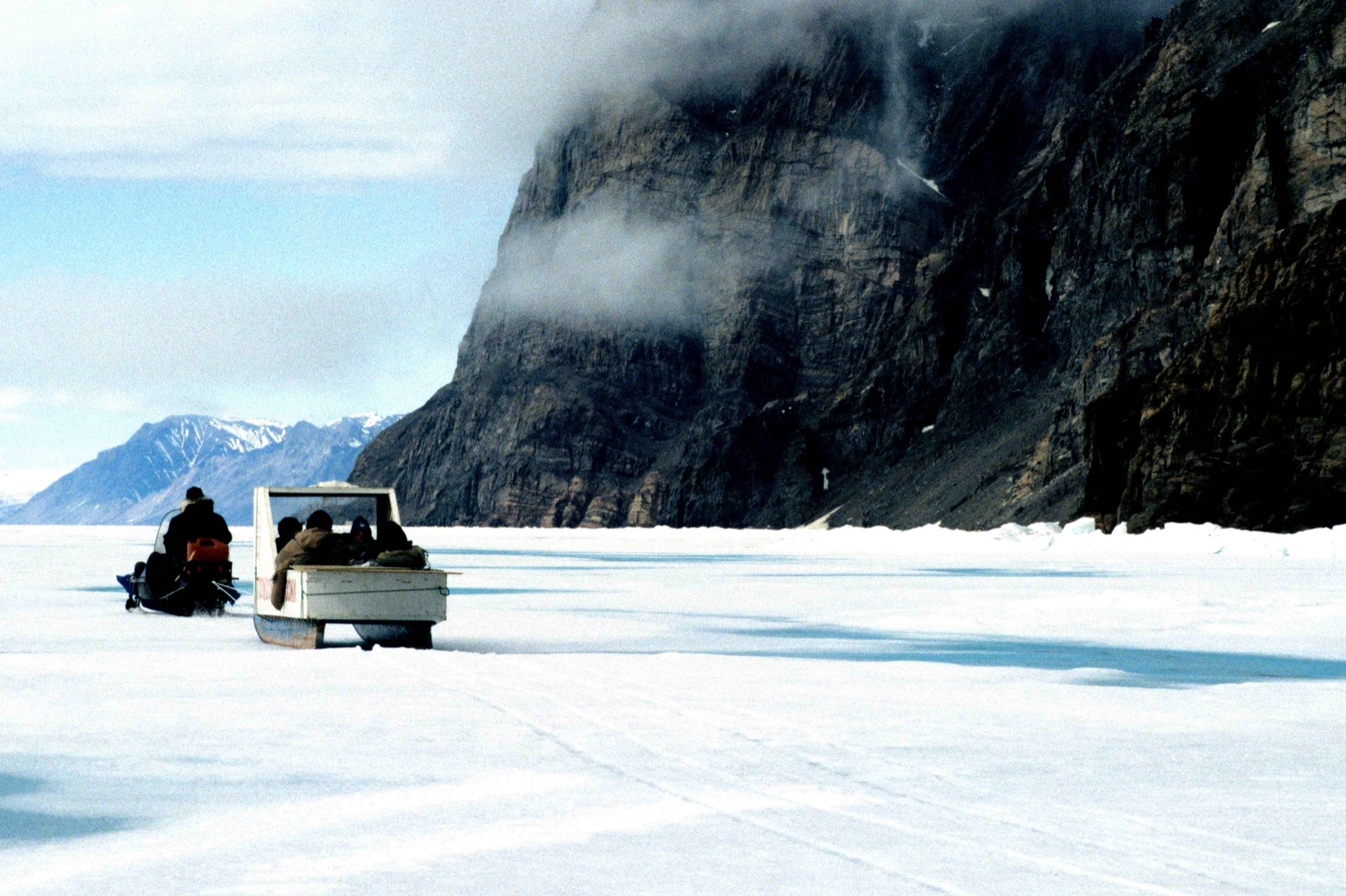 A snowmobile towing a covered sled across flat sea ice below a dark sea cliff on Bylot Island, Nunavut