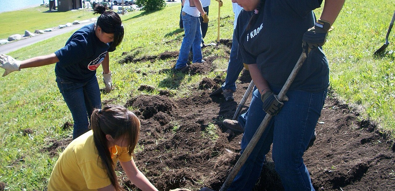 Young people in work gloves digging and planting on a grassy slope beside a lake